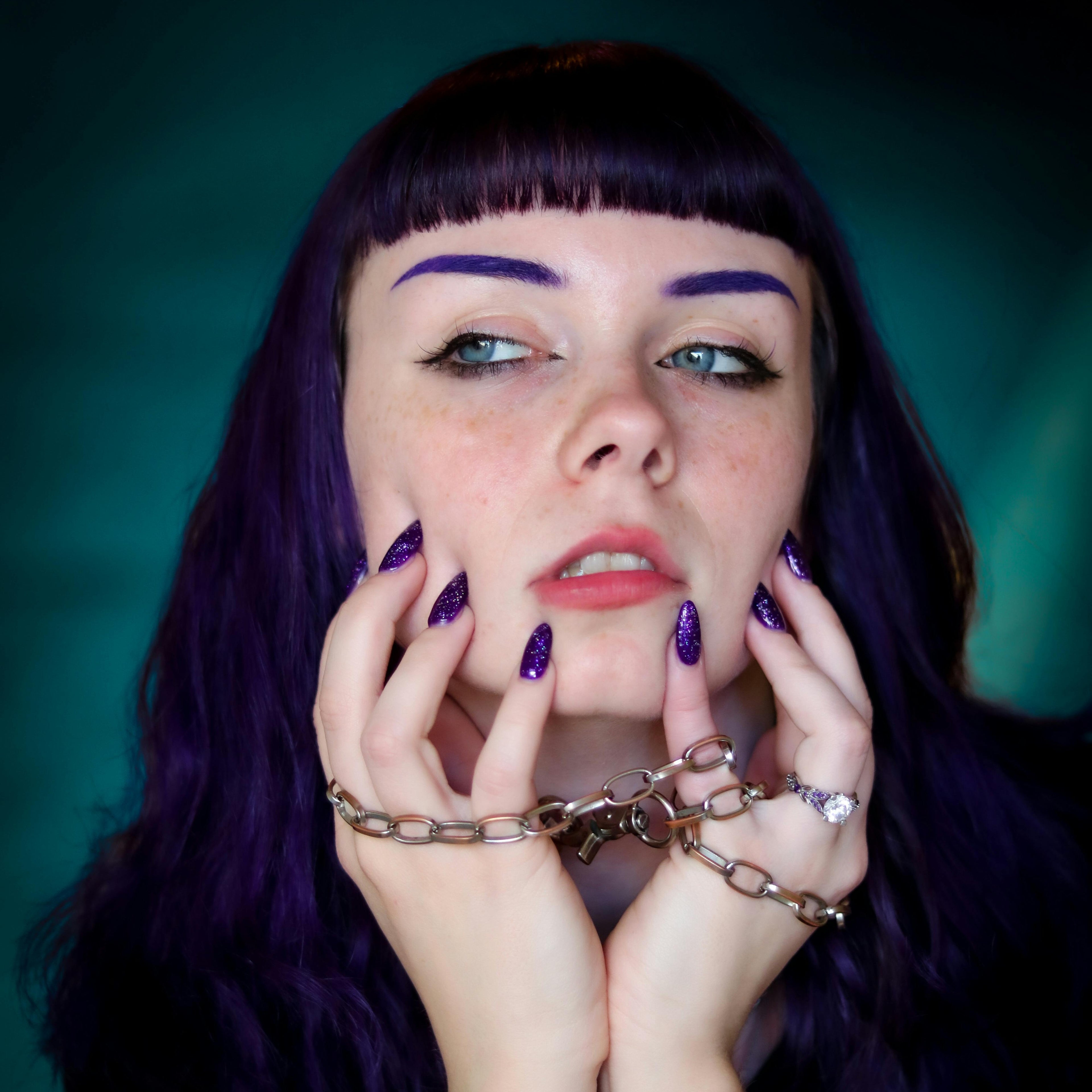 Woman with purple hair and nails holding her hands up to her face against a dark background